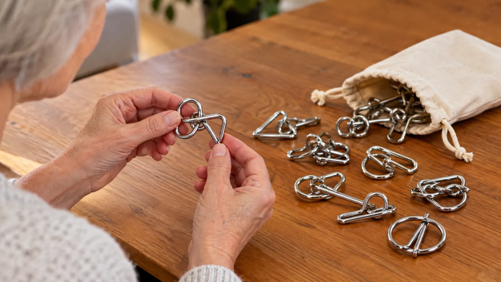 A hand holding a small metal wire puzzle above a worn canvas pouch with other puzzle pieces spilling out onto a wooden table