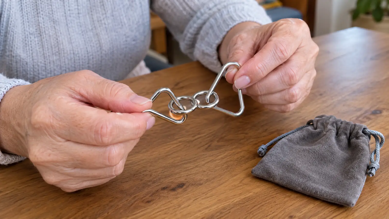 Hands working two linked metal wire pieces over a wooden table with a small drawstring pouch nearby