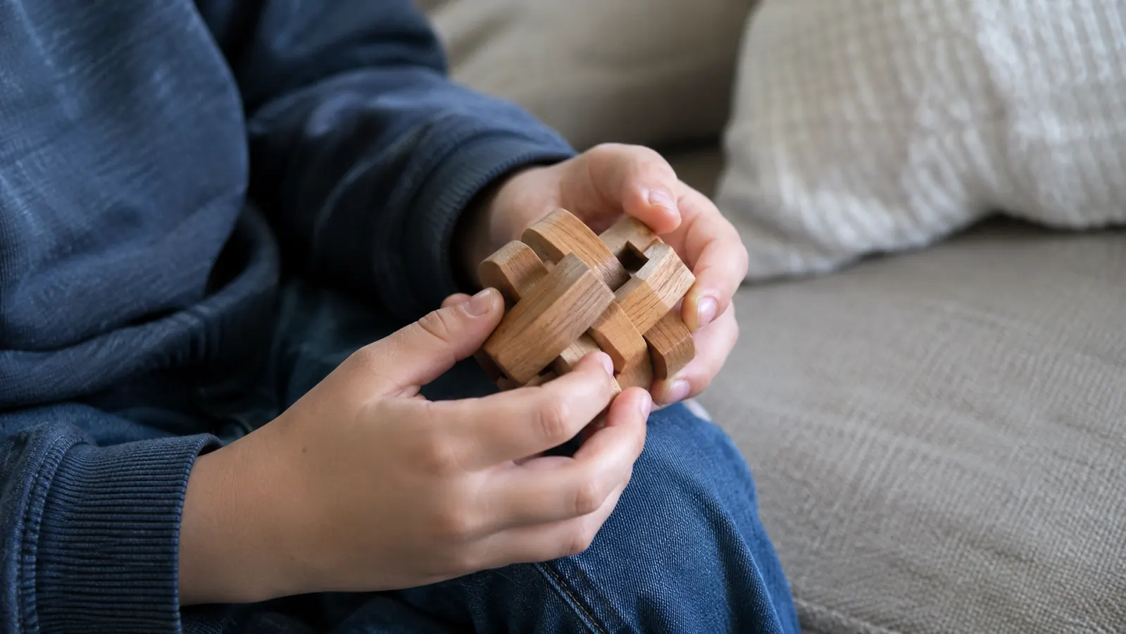 A child's hands working a wooden brain teaser on a couch cushion near a window