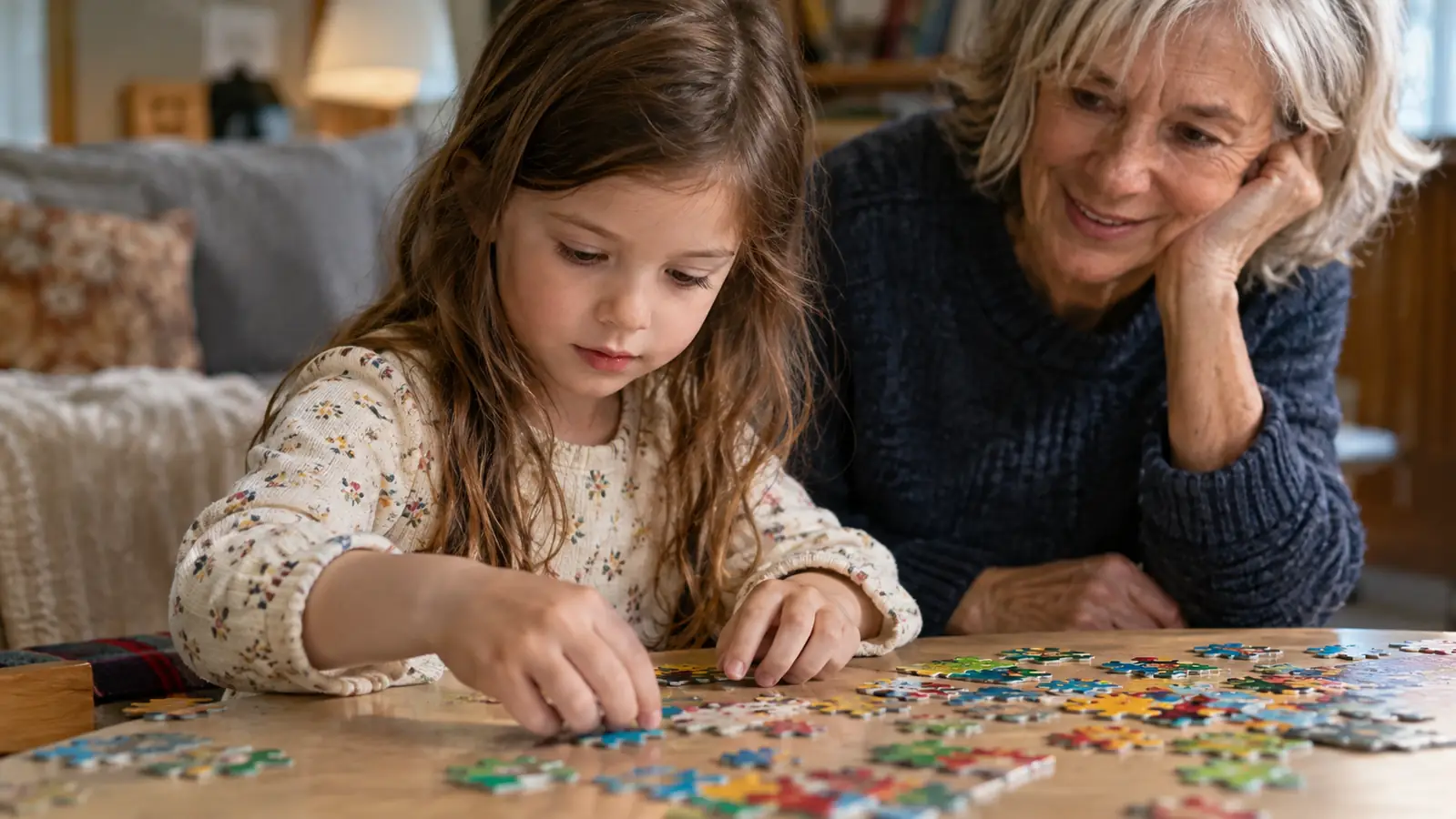 A child's hands working with colorful puzzle pieces on a low table, a grandparent watching nearby